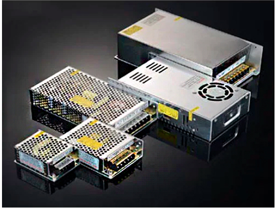 grupo de fontes de alimentação ou drivers metálicos, de formato retangular, dispostos sobre uma superfície escura e reflexiva. Os dispositivos possuem um acabamento prateado ou cinza. Têm tampas ou grades vazadas para ventilação. Há terminais de conexão expostos em uma das extremidades.