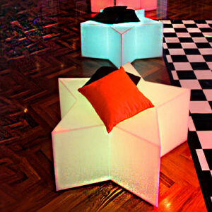 Trata-se de uma foto de dois pufes ou bancos iluminados para festas, no formato de estrelas de cinco pontas. O objeto em primeiro plano é um puff estrela branco translúcido, emitindo um brilho suave com tons avermelhados na base, sobre um chão de madeira. Há uma almofada macia e quadrada de cor laranja vibrante em cima dele. Mais ao fundo, há outro puff estrela similar, com uma almofada escura. No lado direito, vê-se parte de um tapete com padrão xadrez preto e branco.