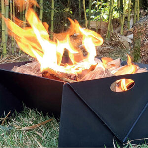A imagem mostra uma fogueira acesa dentro de uma lareira portátil de metal preto, com formato angular. As chamas são grandes, alaranjadas e vibrantes, consumindo pedaços de madeira. A lareira está sobre a grama curta de um jardim. Ao fundo, uma parede densa de bambus altos cria um ambiente natural. A cena sugere calor, luz e um momento de lazer ao ar livre.