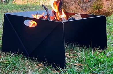 A imagem mostra uma lareira de chão em formato quadrado, feita de metal preto. No centro há fogo aceso com lenhas queimando. A lareira está posicionada sobre a grama, em um ambiente externo, com uma pedra grande ao fundo.
