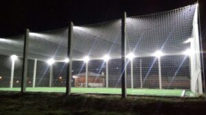 A imagem noturna mostra uma quadra esportiva (gramado sintético verde) cercada por grandes redes de proteção presas a postes verticais escuros. Múltiplos holofotes brancos e brilhantes estão fixados no topo da estrutura, iluminando intensamente a quadra, permitindo o uso noturno. Há uma pequena construção ao fundo.