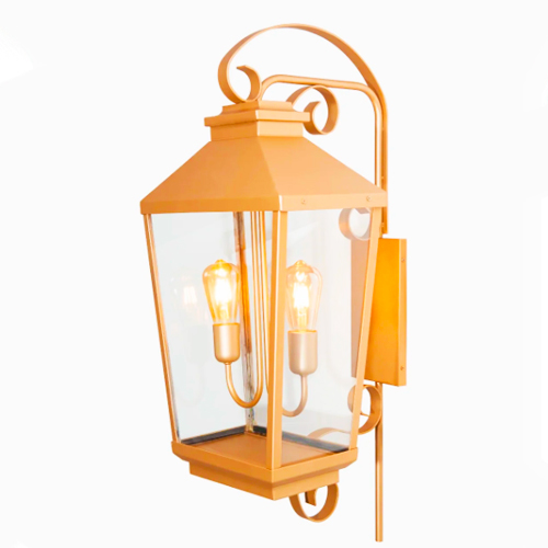 Arandela clássica de metal dourado fixada na parede. Tem formato de lanterna antiga com vidros transparentes, protegendo duas lâmpadas de filamento acesas. O design traz detalhes em curvas (arabescos) no topo e na base, transmitindo um estilo elegante e colonial.