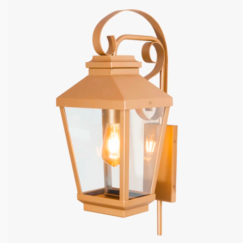 Arandela de parede estilo colonial em metal dourado fosco. Possui formato de lanterna clássica com quatro faces de vidro transparente, revelando uma lâmpada acesa no centro. O topo é decorado com hastes curvas em arabesco, presas a uma base retangular vertical.