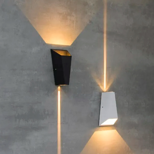 A imagem mostra duas luminárias de parede (arandelas) modernas em formato trapezoidal, uma preta e uma branca. Elas criam um efeito de luz geométrica: de um lado sai um feixe largo e triangular; do outro, um facho fino e vertical, como uma linha de luz sobre a parede