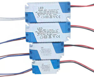 A imagem mostra quatro fontes (drivers) para LED empilhadas. São caixas retangulares, brancas e azuis. Têm especificações impressas, como tensão de entrada ("Input: 110V−220V"), saída ("Output: 45V−84V / 280−300mA"), e modelos como "ABT-15-24X1W". Fios elétricos coloridos (vermelho, preto, branco/azul) saem delas.